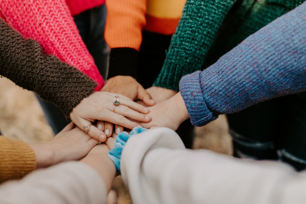 Close up of hands on top of one another in a supportive team huddle. They are wearing bright sweaters and there is a feeling of camaraderie. 