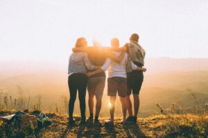 Group of four people standing on a cliff a sunset with their arms around each others' shoulders.