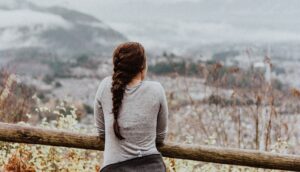 Woman with braid, presumably with ADHD or an Adderall Addiction is looking out over a wooden fence at a mountain range.