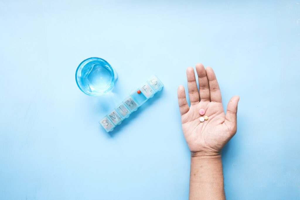 On a light blue background, a person's hand is holding three small antidepressant pills next to a weekly pill organizer and a glass of water. 