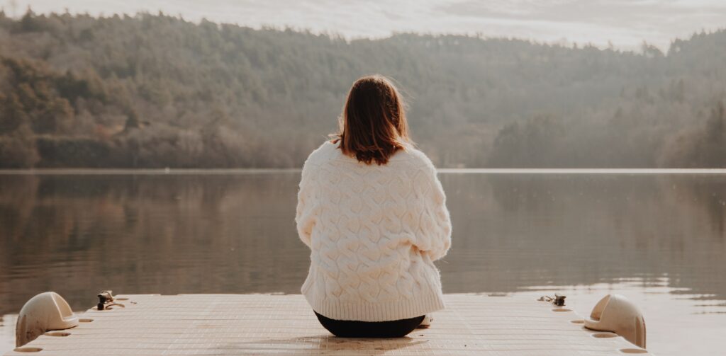 A woman sitting in a white cardigan looking out at a lake wondering how long percocets stay in your system. 