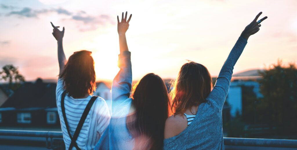 Three friends are standing facing a sunset with their arms in the air, presumably having fun while staying sober at the Newport Beach Film Festival.