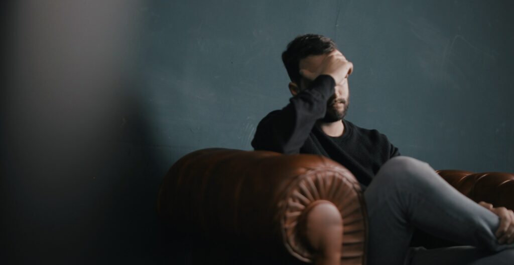 A man is sitting on a leather couch with his hand on his head to represent the symptoms of benzodiazepine addiction (benzo addiction). 
